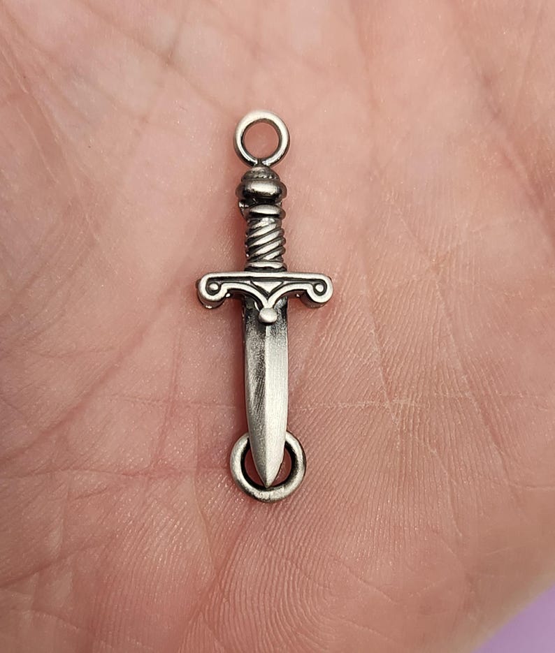May include: Silver-toned sword charm with a detailed design. The charm has a loop at the top for attaching to a necklace or bracelet.