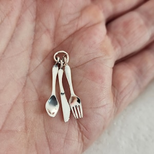 May include: A silver charm featuring miniature cutlery: a spoon, knife, and fork. The charm is designed with a small loop for attachment. The cutlery is highly detailed, reflecting light.