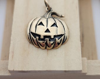 May include: A bronze-colored pumpkin pendant with a carved jack-o'-lantern face. The pendant has triangular eyes, a triangular nose, and a wide, toothy grin. The pumpkin is suspended from a small loop at the top. The pendant is displayed on a light-colored wooden surface.