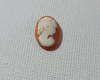 May include: A small, oval-shaped cameo with a white cameo of a woman's profile set against a translucent orange background.
