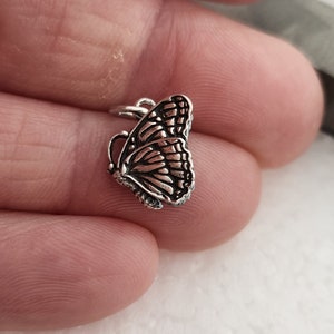 May include: A small, detailed silver butterfly charm with intricate wing patterns. The charm has a loop at the top for attaching to a necklace or bracelet. The butterfly's wings are spread, showcasing the delicate design.