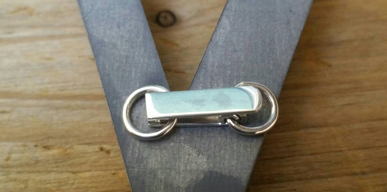 Peut inclure: Gros plan sur un fermoir en m&eacute;tal argent&eacute; avec deux anneaux circulaires et une pi&egrave;ce centrale rectangulaire bleu clair. Le fermoir est fix&eacute; &agrave; une surface en bois grise, avec un fond en bois.