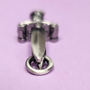 May include: A silver-toned sword charm with a ring at the bottom. The sword is detailed with a cross guard and a pointed tip.