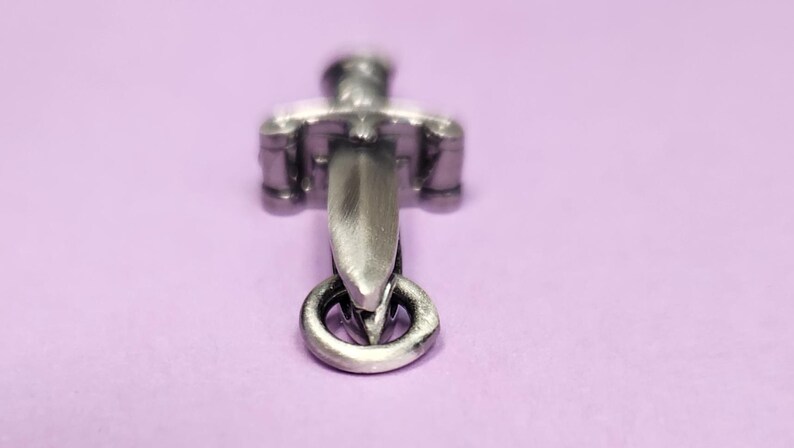 May include: A silver-toned sword charm with a ring at the bottom. The sword is detailed with a cross guard and a pointed tip.