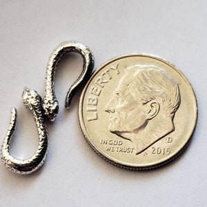 Puede incluir: Un amuleto de serpiente de plata con una superficie texturizada. El amuleto es aproximadamente del tama&ntilde;o de una moneda de diez centavos.