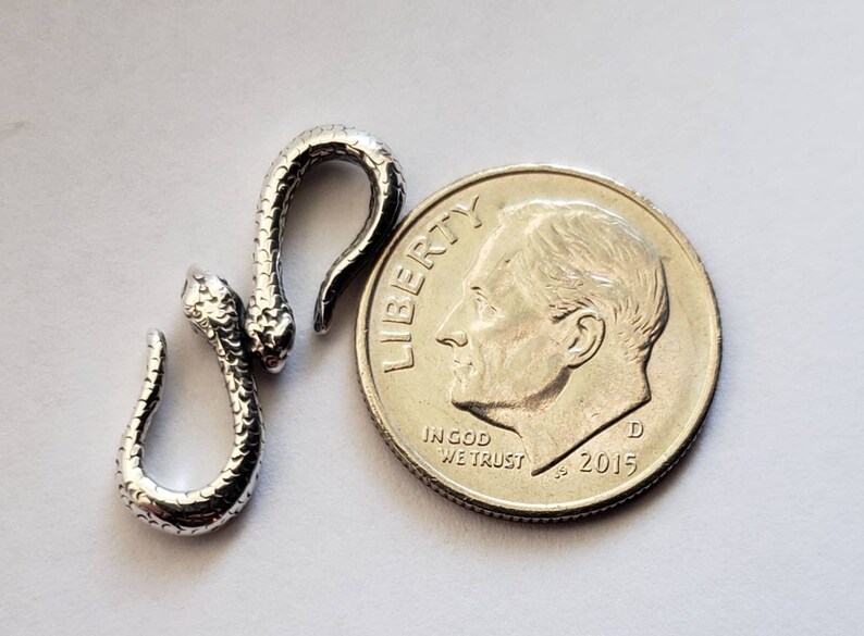 Puede incluir: Un amuleto de serpiente de plata con una superficie texturizada. El amuleto es aproximadamente del tama&ntilde;o de una moneda de diez centavos.