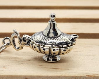 May include: A silver-colored charm in the shape of a genie lamp. The lamp has a detailed design with a small handle and a decorative lid. The charm is resting on a light brown wooden surface.