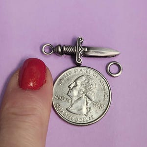 May include: Silver-toned sword charm with a loop for attaching to a necklace or bracelet. The charm is about the size of a US quarter.