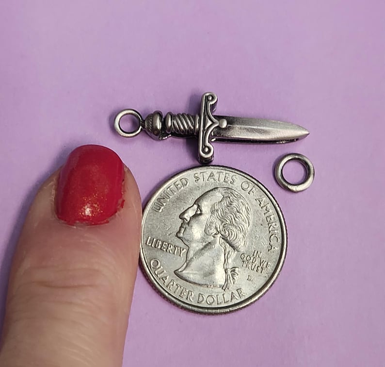 May include: Silver-toned sword charm with a loop for attaching to a necklace or bracelet. The charm is about the size of a US quarter.