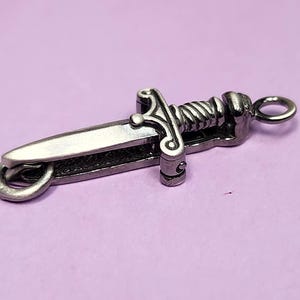 May include: A silver-toned dagger charm with a detailed design and a ring for attaching to a chain or bracelet.