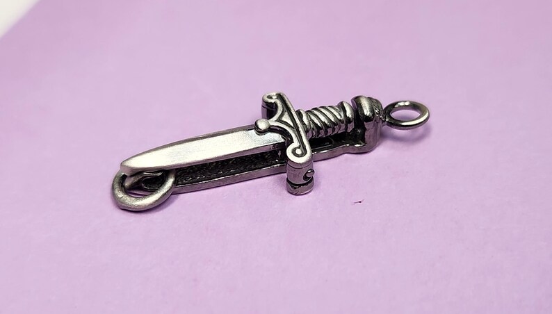 May include: A silver-toned dagger charm with a detailed design and a ring for attaching to a chain or bracelet.