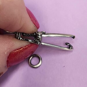 May include: Silver-toned metal clasp with a spring-loaded mechanism. The clasp is open and ready to be attached to a chain or other jewelry.