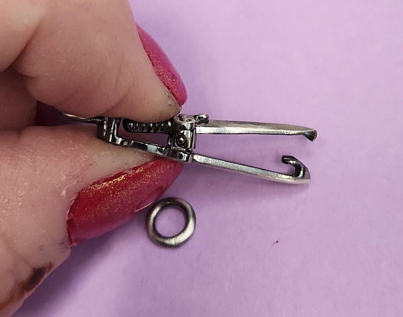 May include: Silver-toned metal clasp with a spring-loaded mechanism. The clasp is open and ready to be attached to a chain or other jewelry.