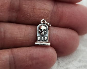 May include: A small silver-colored charm shaped like a tombstone, featuring a raised skull design at the top and the letters RIP embossed clearly below it. The charm includes a small circular jump ring attached to the top for hanging purposes.