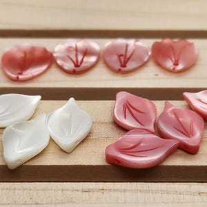 May include: An assortment of shell-shaped beads in shades of pink and white, arranged on a wooden surface. The beads are in the shape of petals and leaves, with delicate veining details.