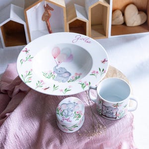 Peut inclure: Ensemble de vaisselle en céramique blanche comprenant un bol, une tasse et une boîte à bijoux. Motif éléphant et ballons, avec le nom "Jolie" en cursive. Fleurs roses et feuilles vertes ornent le bol et la tasse. La boîte est ronde.