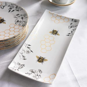 Può includere: Vassoio da portata in ceramica bianca con design a forma di ape e favo. Il vassoio rettangolare ha bordi leggermente curvi. Altri articoli includono piatti, tazze e piattini coordinati, tutti con finiture dorate e motivi a forma di ape.