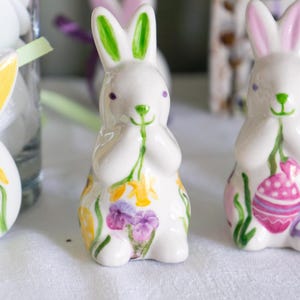 Puede incluir: Dos figuritas de conejo de cerámica blanca con detalles florales pintados. Un conejo sostiene un tallo verde con flores amarillas y el otro sostiene un tallo verde con un huevo de Pascua rosa y morado.