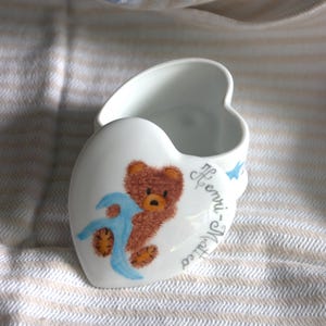 Könnte beinhalten: Eine weiße herzförmige Schmuckdose aus Porzellan mit einem Deckel, der einen braunen Teddybären mit einer blauen Decke zeigt. Der Name Henri-Matteo ist in Schreibschrift auf den Deckel geschrieben. Die Dose hat eine glatte, glänzende Oberfläche.