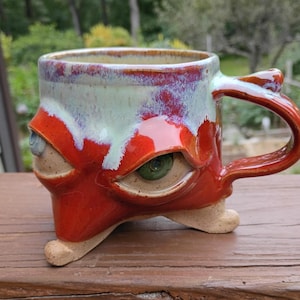 May include: A ceramic mug with a glossy red glaze and a pale green drip pattern at the rim. The mug features two sculpted, realistic eyes embedded in the sides and stands on three small, beige, foot-shaped protrusions at the base.
