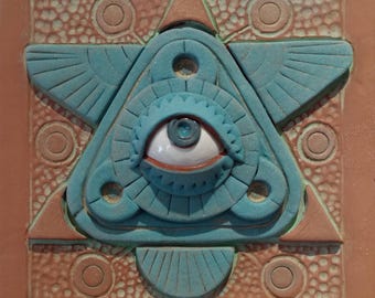 May include: A square, ceramic tile featuring a central turquoise eye within a triangle, surrounded by stylized wings and geometric shapes. The tile has a brown background with circular and triangular accents, creating a textured, artistic design.