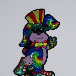 Puede incluir: Una pegatina holográfica colorida de un conejo de dibujos animados que lleva un sombrero de tío Sam rojo, blanco y azul, gafas de sol y una pajarita. El conejo está montando una patineta con estrellas y rayas.