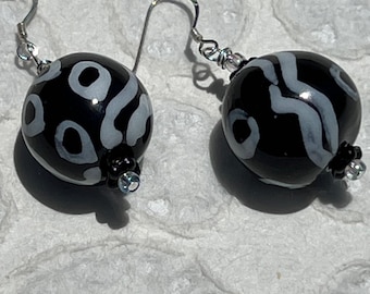 May include: A pair of dangle earrings featuring spherical black glass beads with white abstract patterns. Each bead is accented with a small black flower-shaped spacer and a tiny clear bead at the base, suspended from silver-toned metal ear wires.