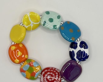 May include: A stretch bracelet composed of nine smooth, rounded, multi-colored ceramic beads. The beads feature various patterns including solid colors, polka dots, swirls, and abstract shapes in yellow, orange, red, purple, blue, and green.