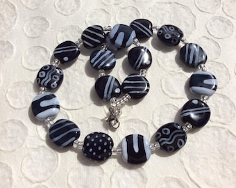 May include: A necklace featuring large, oval beads in black and light gray. The beads are strung together with a silver clasp and small clear beads. The black beads have various white patterns, including stripes and dots.