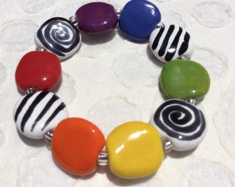 May include: A stretch bracelet featuring smooth, rounded, pebble-shaped beads in a variety of colors including red, orange, yellow, green, blue, and purple, interspersed with white beads decorated with black spiral or striped patterns and small silver spacers.