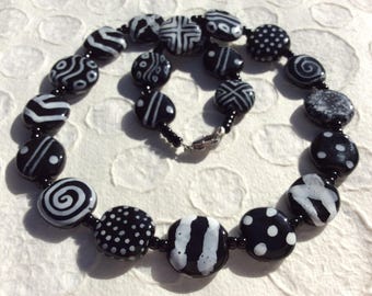 May include: A black and white beaded necklace with various patterns. The necklace features round beads with designs like polka dots, swirls, and geometric shapes. The beads are strung together with smaller black beads and a silver clasp.