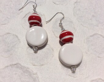 May include: A pair of dangle earrings featuring silver-toned hooks, each with a round red bead accented by two horizontal white stripes, suspended above a larger, smooth, flat white circular disc bead with a small clear bead at the base.