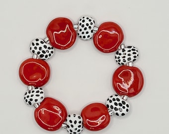 May include: A circular bracelet composed of alternating smooth, glossy red ceramic beads and smaller, round white beads featuring a black spotted pattern. The beads are strung together with small silver-toned spacers between each individual bead.