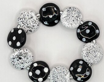 May include: A circular bracelet composed of alternating glass beads. The beads feature either a glossy black finish with white polka dots and thin silver-toned lines or a textured white and black speckled finish, all strung together on a flexible cord.
