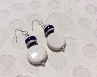 May include: A pair of drop earrings featuring a round, white, crackle-textured bead suspended below a smaller, spherical, dark blue bead with horizontal white stripes. The earrings are attached to silver-toned metal hooks for pierced ears.
