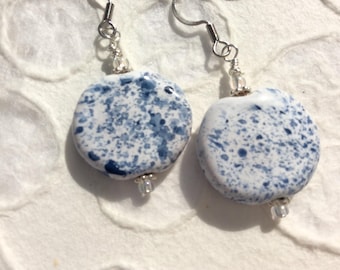 May include: A pair of handmade earrings featuring round, speckled ceramic beads in white and blue. The earrings have silver-tone hooks and small clear beads at the bottom of each earring. The speckled design gives the earrings a unique, artistic look.