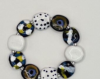 May include: A bracelet composed of ten irregular, smooth, glossy beads. The beads feature varied designs including white with black dots, solid white, and abstract patterns in navy blue, yellow, white, and metallic gold, separated by small silver spacers.