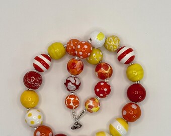 May include: A beaded necklace featuring a variety of round, colorful beads in shades of red, orange, yellow, and white. The beads display diverse patterns including solid colors, polka dots, and stripes, all strung together on a clear cord with a metal clasp.