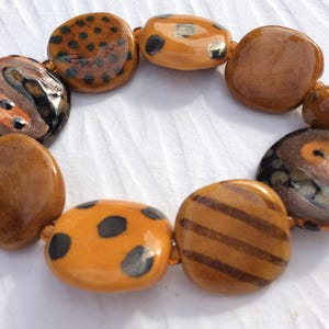 May include: A handmade bracelet featuring large, irregularly shaped beads in shades of brown and orange. Some beads have black spots or stripes, while others are solid. The bracelet is strung on a neutral-colored cord.