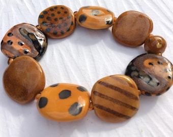 May include: A handmade bracelet featuring large, irregularly shaped beads in shades of brown and orange. Some beads have black spots or stripes, while others are solid. The bracelet is strung on a neutral-colored cord.