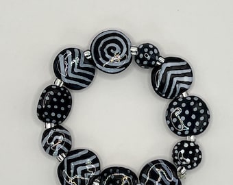 May include: A circular bracelet composed of black glass beads featuring hand-painted white patterns including polka dots, chevrons, and spirals. The beads are separated by small silver-toned metallic spacers, creating a high-contrast monochrome design.