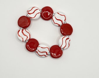 May include: A circular bracelet composed of alternating round beads. The beads consist of solid glossy red discs and white discs featuring wavy red painted lines. The beads are strung together in a repeating pattern to form a continuous loop.