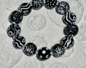 May include: A circular bracelet composed of fourteen round beads in a black and white color scheme. The beads feature various patterns including white polka dots, abstract white swirls, and speckled white textures on a glossy black base.