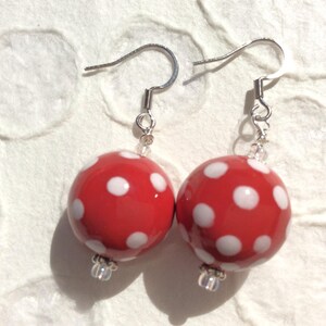 May include: A pair of red, round earrings with white polka dots. The earrings have silver-colored hooks and small clear beads. The earrings are a fun, playful accessory.