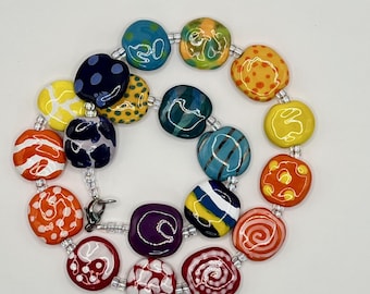 May include: A necklace featuring a variety of colorful, flat, rounded beads with diverse patterns including dots, swirls, and stripes in shades of red, orange, yellow, green, blue, and purple, connected by small clear spacer beads and a metal clasp.