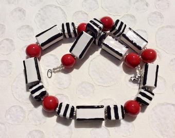 May include: A necklace featuring alternating red spherical beads and rectangular white beads with black stripes. The necklace is strung on a silver chain and has a silver clasp. The beads are approximately 1 cm in diameter.