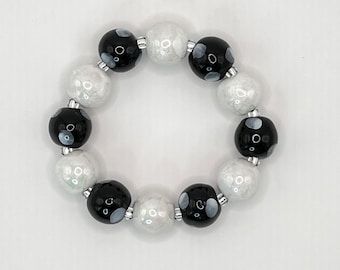 May include: A circular bracelet composed of alternating spherical beads. The beads consist of glossy black spheres featuring small white circular patterns and textured, iridescent white spheres. Small silver-toned spacers are placed between each bead.