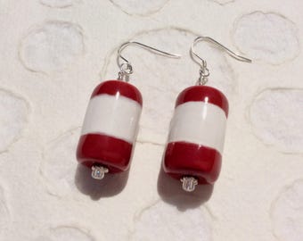 May include: A pair of handmade earrings featuring a cylindrical design with alternating bands of red and white. The earrings have silver-colored hooks and small decorative silver-colored accents at the bottom.