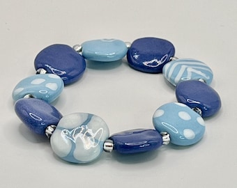 May include: A stretch bracelet composed of smooth, rounded, disc-shaped beads in various shades of blue. The beads feature patterns including white polka dots, stripes, and solid colors, separated by small, reflective silver-toned spacer beads.
