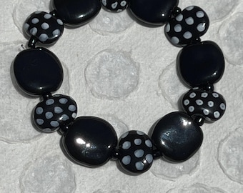 May include: A black beaded bracelet with a mix of solid and polka-dot beads. The bracelet features large, glossy black beads interspersed with smaller beads adorned with white polka dots. The beads are strung together to form a circular shape.